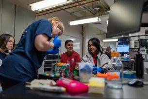 Four students in a lab, with one conducting an experiment while others watch